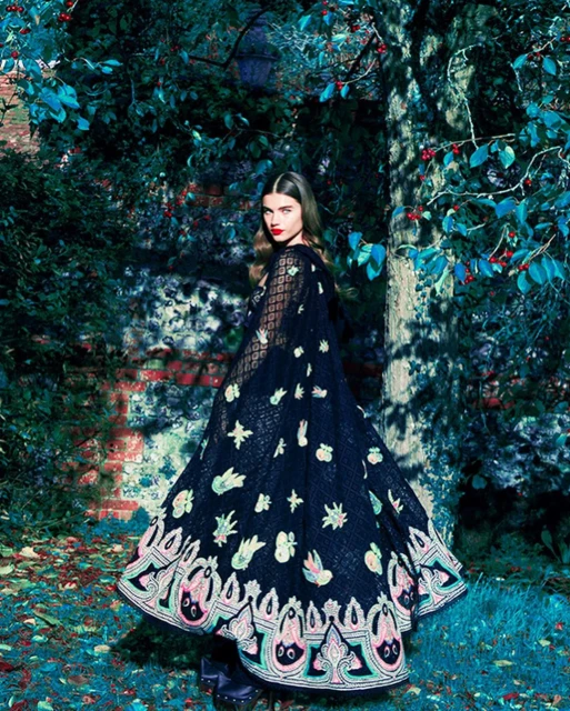 An adult female model poses outdoors wearing a black cape with floral embellishments, standing in a lush garden with a brick wall.