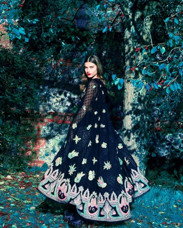 An adult female model poses outdoors wearing a black cape with floral embellishments, standing in a lush garden with a brick wall.