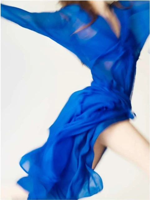 Blurred action shot of a person in motion, wearing a draped royal blue dress against a white background, showcasing movement and fluidity.