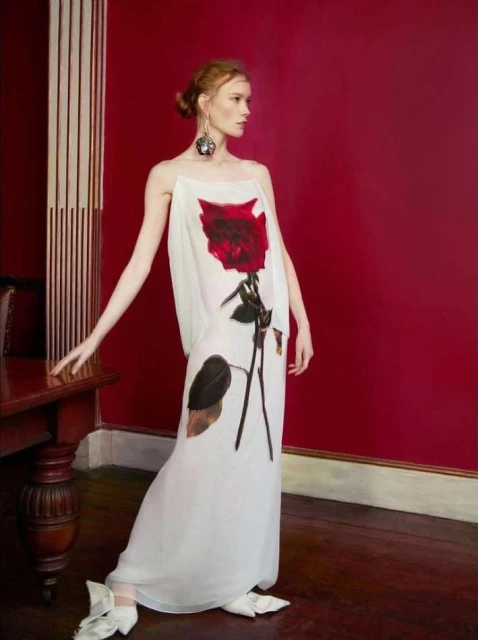 Elegant woman in a white floral dress, posing in a vintage interior setting against a rich red wall. Fashion editorial shot.
