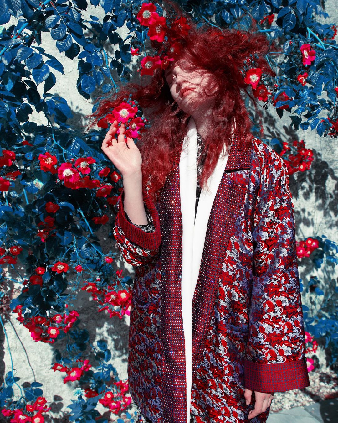 Woman with red hair poses in front of flowering vines wearing a patterned robe with white trim. Red flowers accent the hair.