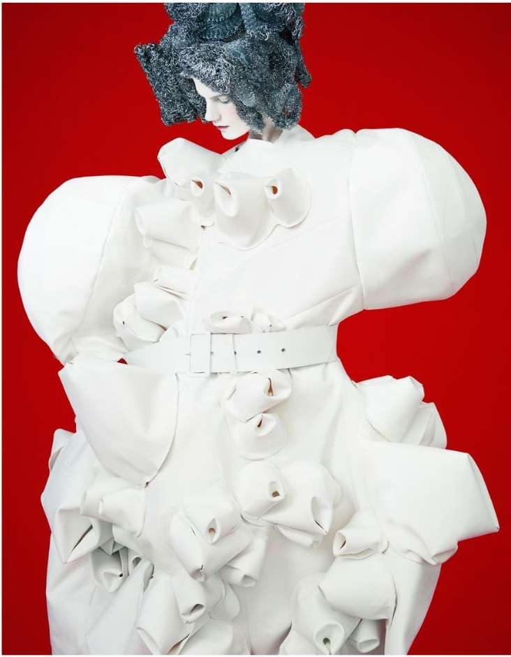 Fashion model in an avant-garde white dress with a ruffled design poses against a vibrant red background for a high-fashion editorial.