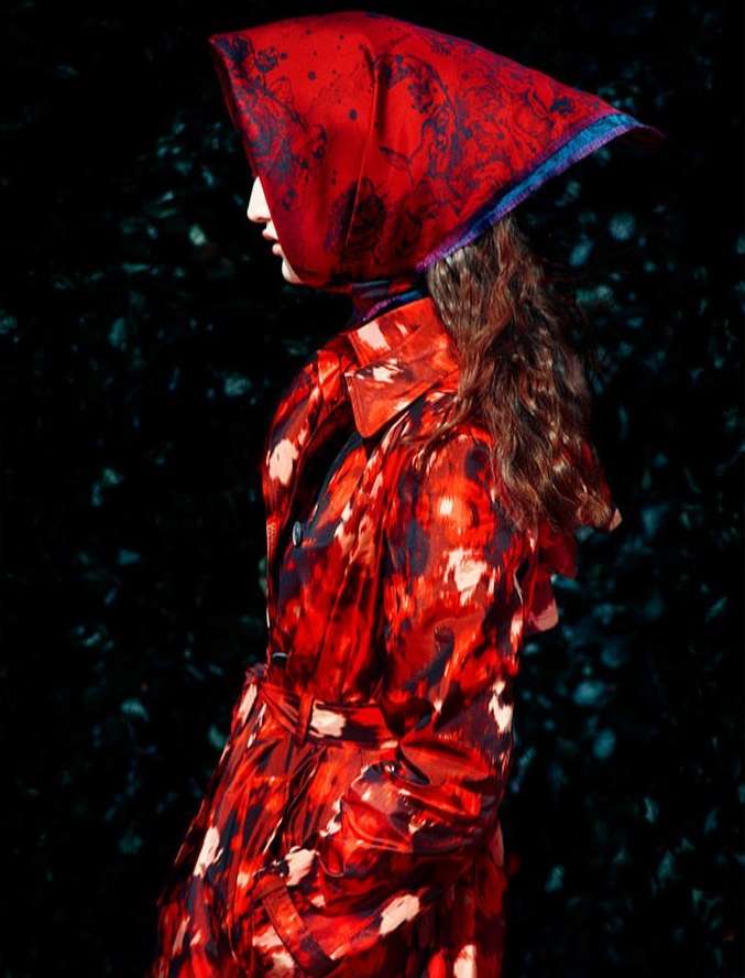 Profile of a woman with long curly hair wearing a vibrant red patterned coat and matching headscarf against a dark background.