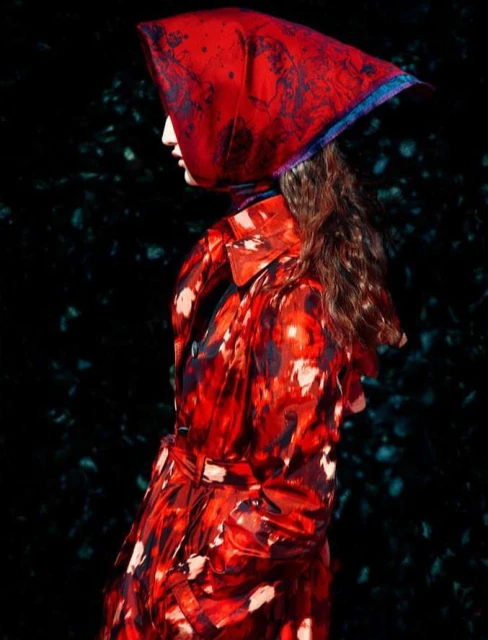 Profile of a woman with long curly hair wearing a vibrant red patterned coat and matching headscarf against a dark background.