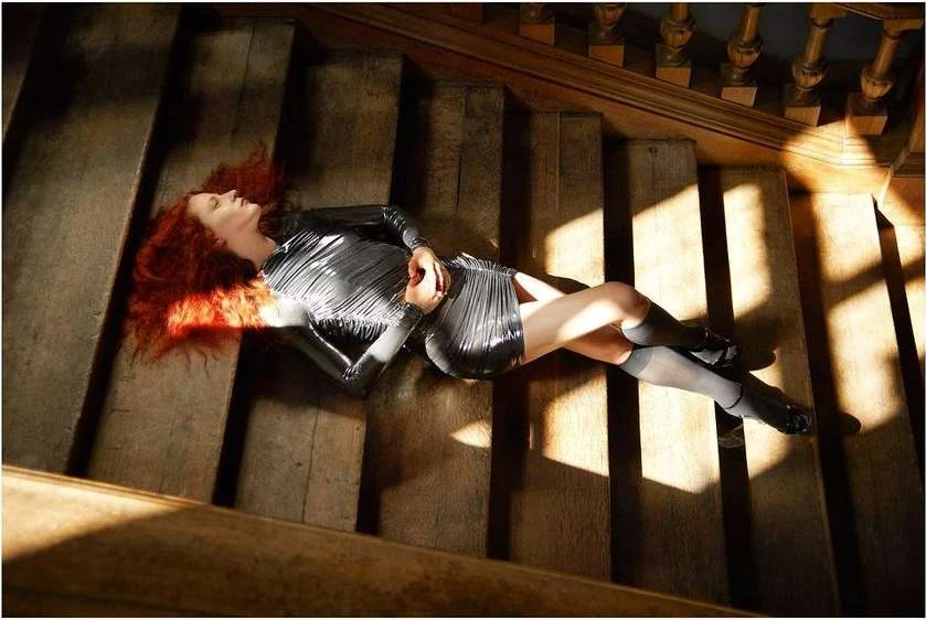 Redhead woman in a silver dress lies on wooden stairs illuminated by sunlight. The scene evokes a sense of mystery and artistry.