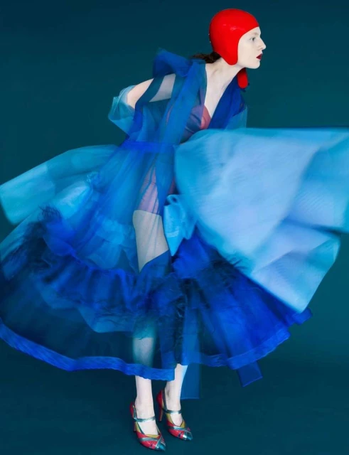 Fashion portrait of a model wearing a blue gown and a red cap against a teal background in a studio with dramatic lighting.