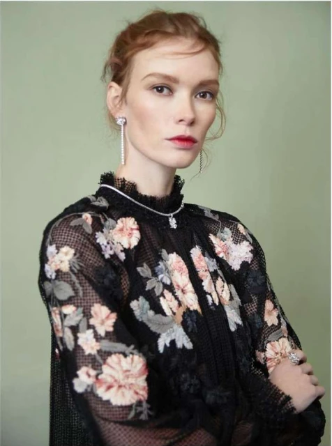Portrait of a redhead woman wearing a black floral-patterned mesh top and jewelry, against a light green backdrop, in a beauty fashion shot.