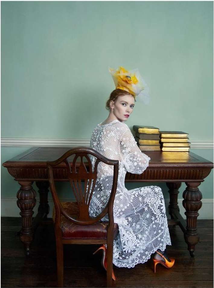 Adult woman in a white lace dress and hat posing in a photo studio while sitting at a table with stacked books and wearing heels.
