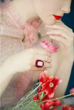 Young woman in pink dress with red lipstick and nail polish holds pink flowers, featuring a large ring with a red gemstone.