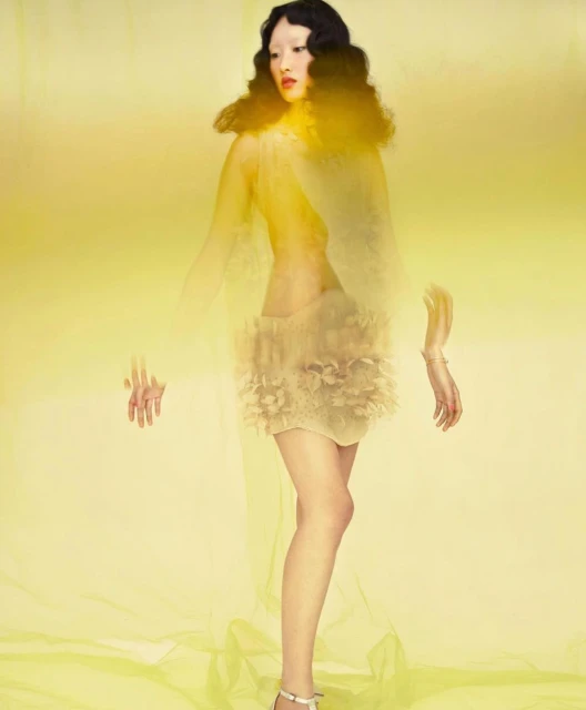 Elegant Asian woman in flowing yellow outfit with diaphanous fabric and decorative skirt poses against a bright yellow background.