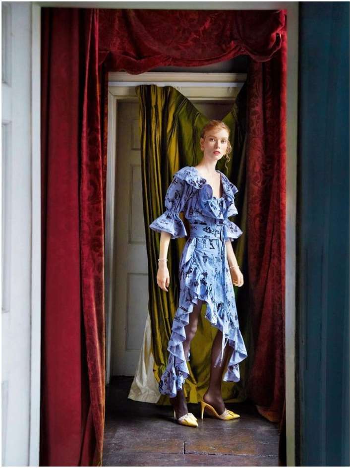 Fashion shot of a red-haired woman in a blue floral dress, yellow heels, and dark stockings, standing in front of a doorway with red curtains.