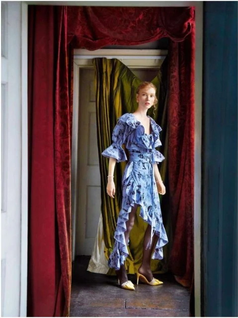 Fashion shot of a red-haired woman in a blue floral dress, yellow heels, and dark stockings, standing in front of a doorway with red curtains.