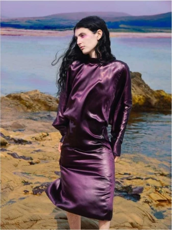 Model in a lustrous purple satin dress standing on rocky terrain near water under a pale sky, photographed by Erik Madigan Heck.