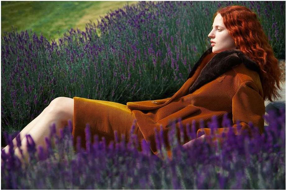 A red-haired woman in a tan corduroy jacket lies amongst purple lavender flowers in a field on a sunny day in an outdoor setting.