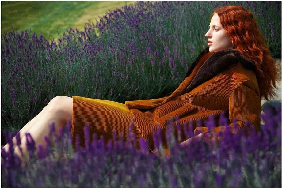 A red-haired woman in a tan corduroy jacket lies amongst purple lavender flowers in a field on a sunny day in an outdoor setting.