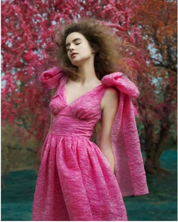 A young woman poses in a pink dress with large bow sleeves against a red-leafed tree backdrop in a fashion editorial-style photograph.