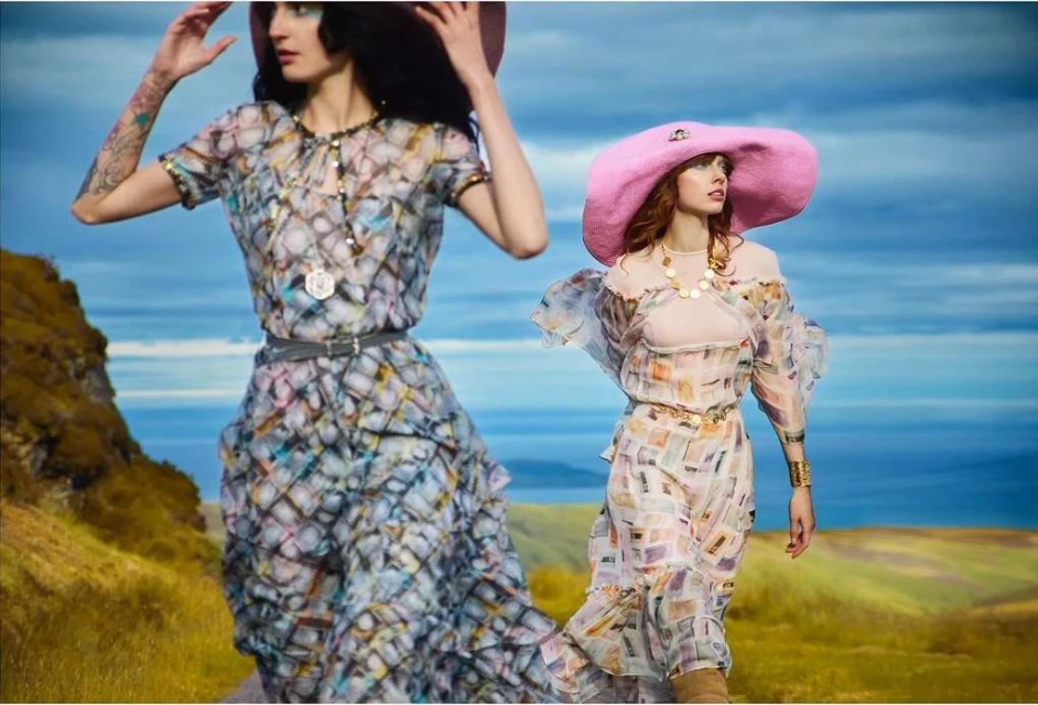 Two women in large hats and flowing dresses pose in a field under a blue sky, showcasing outdoor fashion and elegant style.