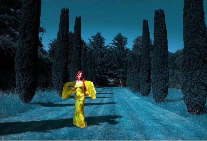 Woman with red hair wearing a vibrant yellow dress standing on a dark blue grassy path flanked by tall cypress trees