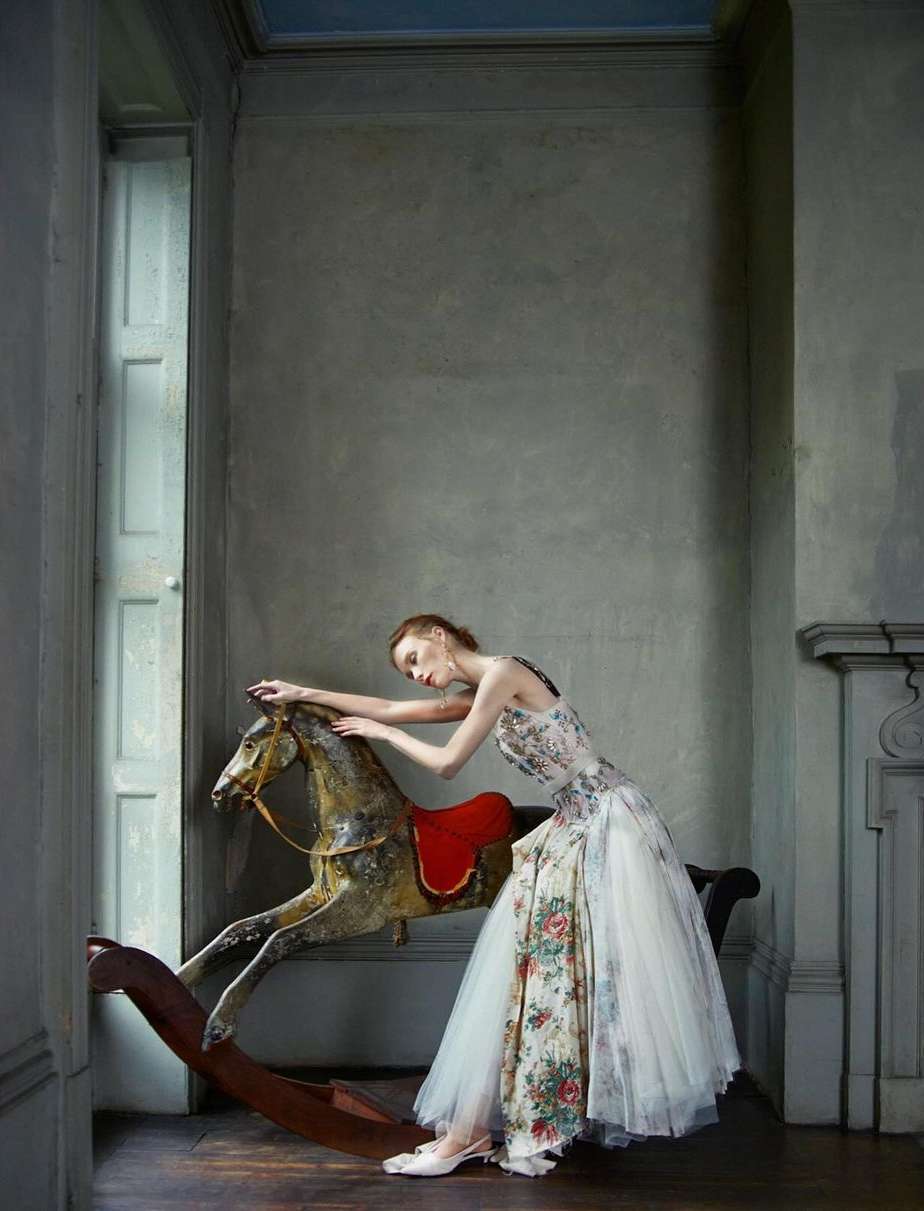 Woman in floral dress poses with antique rocking horse in a room with gray walls and a window, exuding a vintage fashion editorial aesthetic.