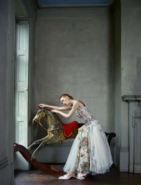 Woman in floral dress poses with antique rocking horse in a room with gray walls and a window, exuding a vintage fashion editorial aesthetic.