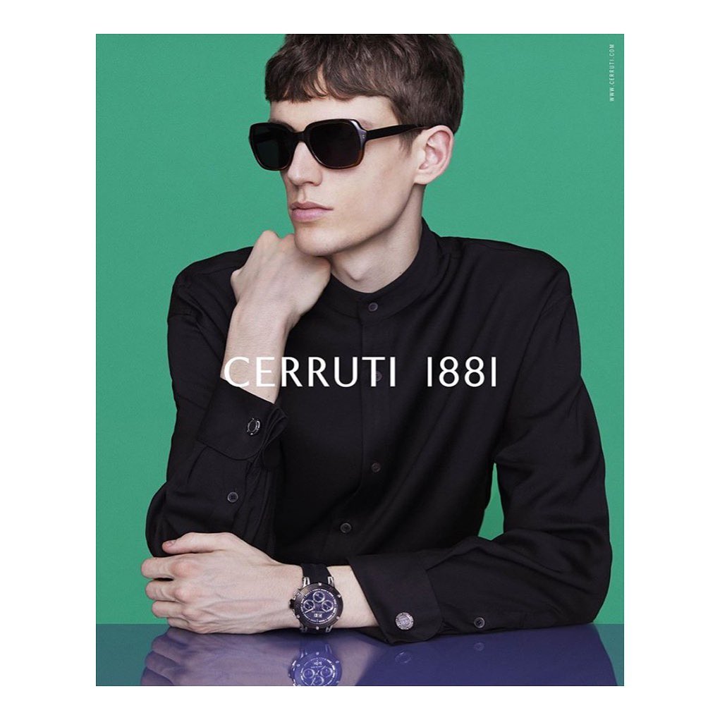 Fashion shot of a young man wearing black sunglasses and a black button-down shirt with a watch against a green and blue background.