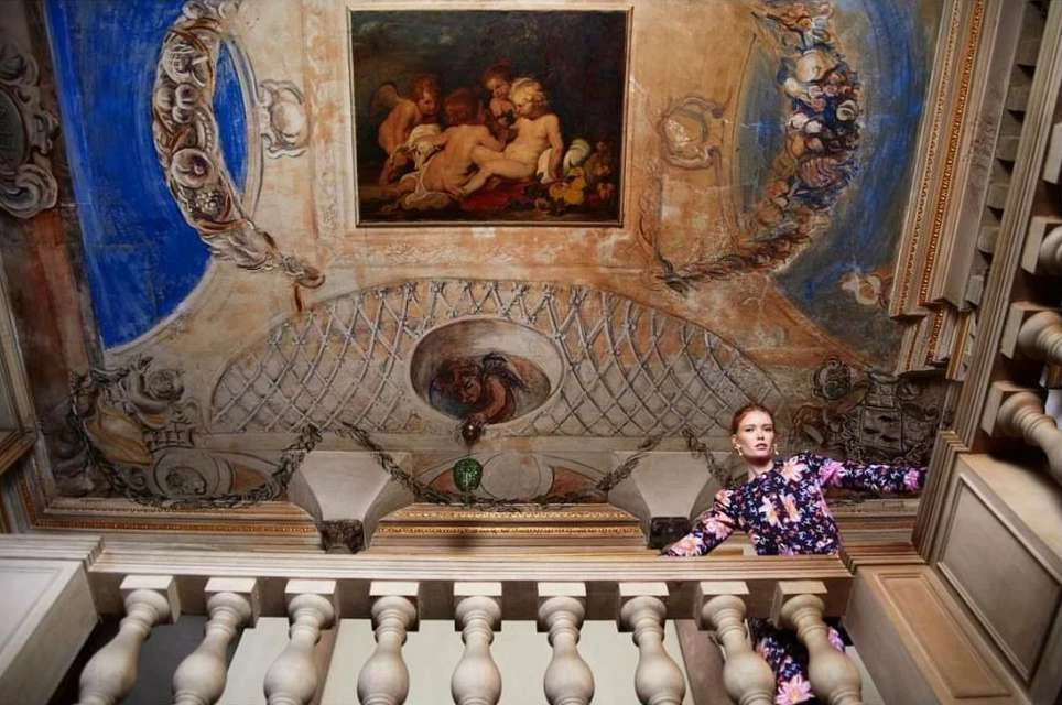 A woman in a floral dress is on a staircase with painted walls and ceiling featuring cherubs, intricate designs, and architectural details.