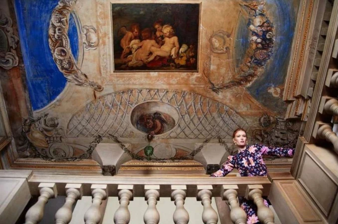 A woman in a floral dress is on a staircase with painted walls and ceiling featuring cherubs, intricate designs, and architectural details.