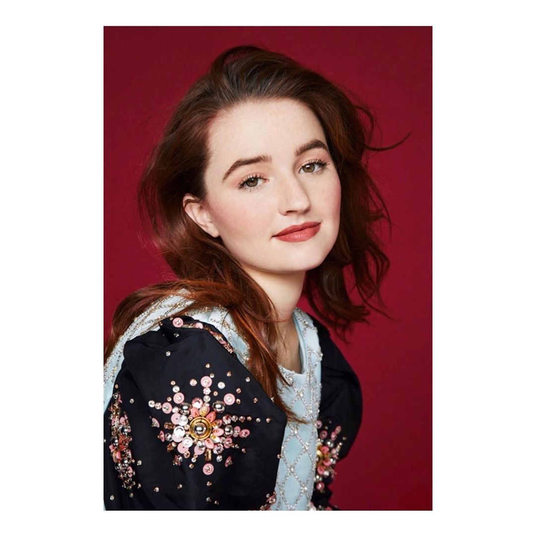 Portrait of a young woman with long auburn hair, wearing a black top with floral embellishments, against a dark red backdrop.
