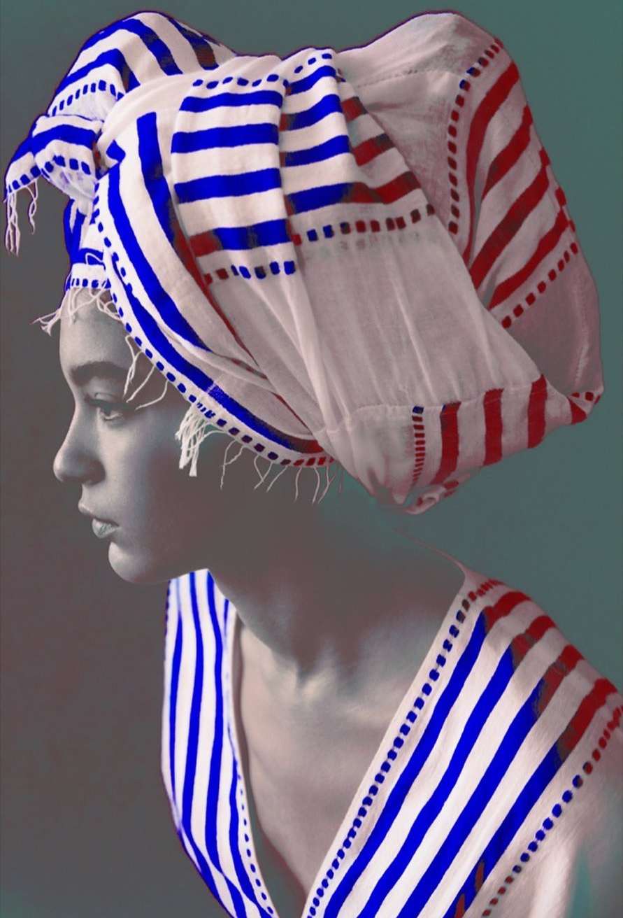A woman in a white and blue striped turban and matching dress looks to the side in a studio shot.
