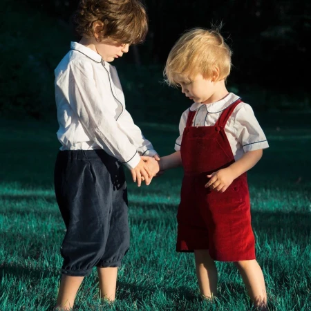 Two young boys holding hands standing on green grass, wearing white shirts and dark trousers and red overalls.