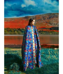 Model wearing a long blue and red floral patterned cape standing on a grassy shore overlooking a body of water and reddish hills.