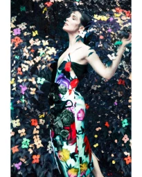 Model in a colourful floral print dress posing against a dense background of dark leaves and vibrant flowers.