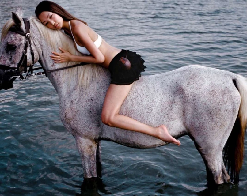 A young woman wearing a white bikini top and dark shorts, is lying on the back of a white horse that stands in the water.
