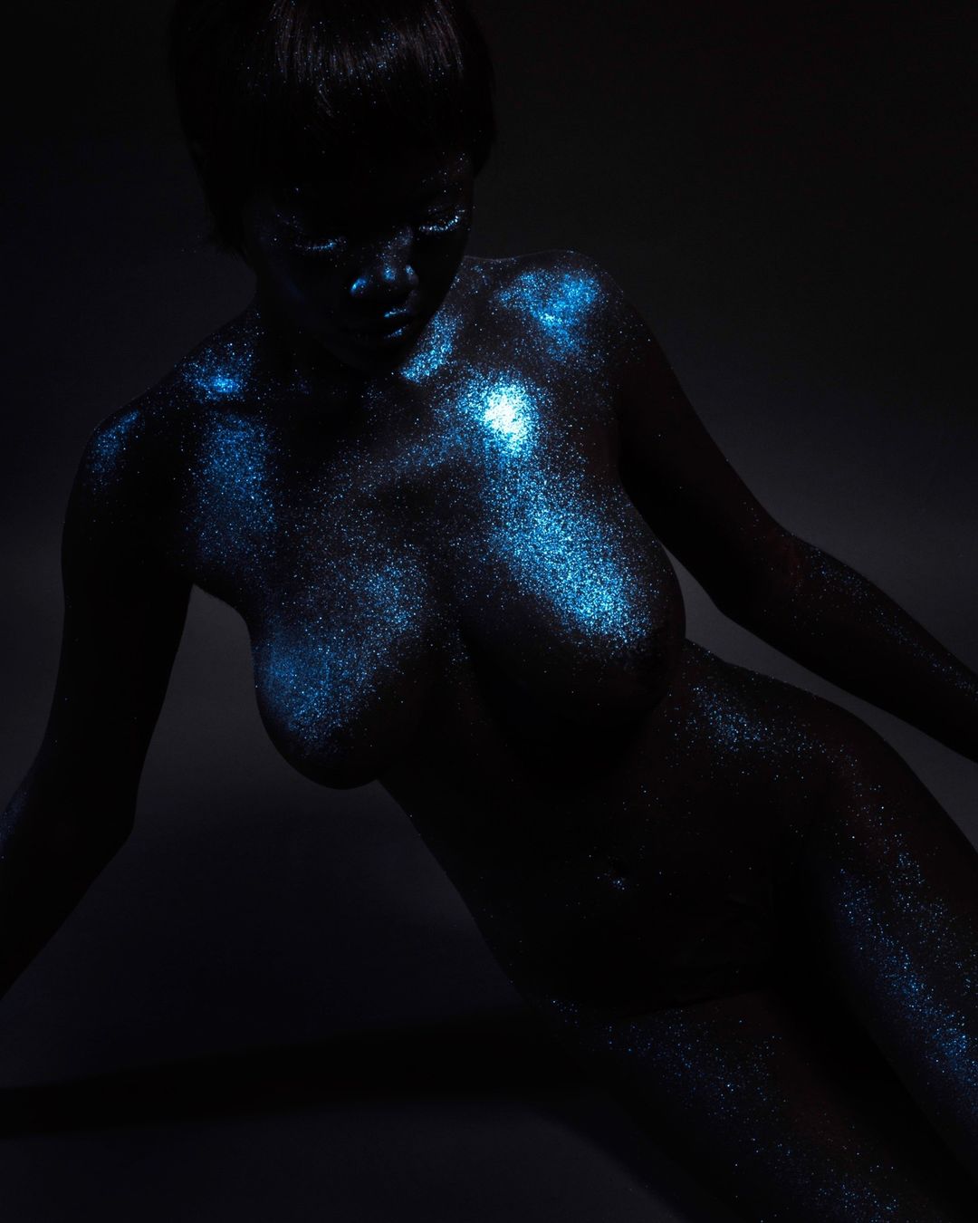 Striking image of a nude adult woman with blue glitter art on her body, set against a dark gray background in a photo studio.
