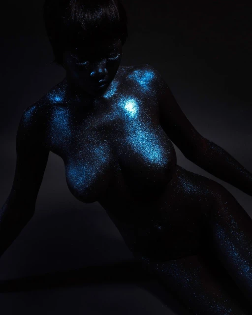 Striking image of a nude adult woman with blue glitter art on her body, set against a dark gray background in a photo studio.