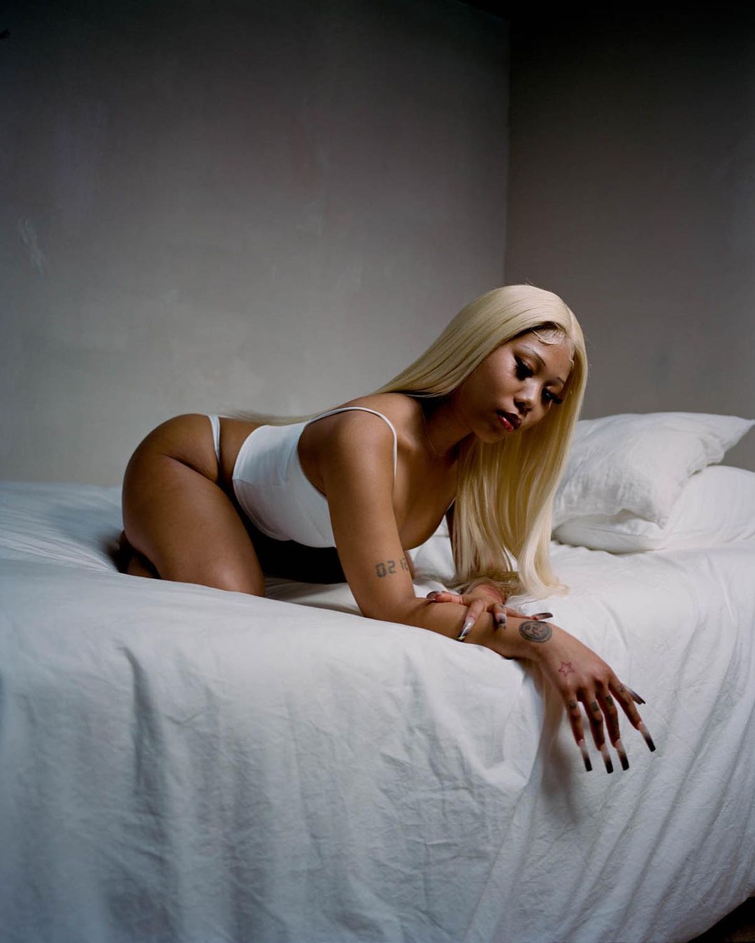 Woman with blonde hair in white lingerie poses on a white bed in an indoor setting with a blank wall background.