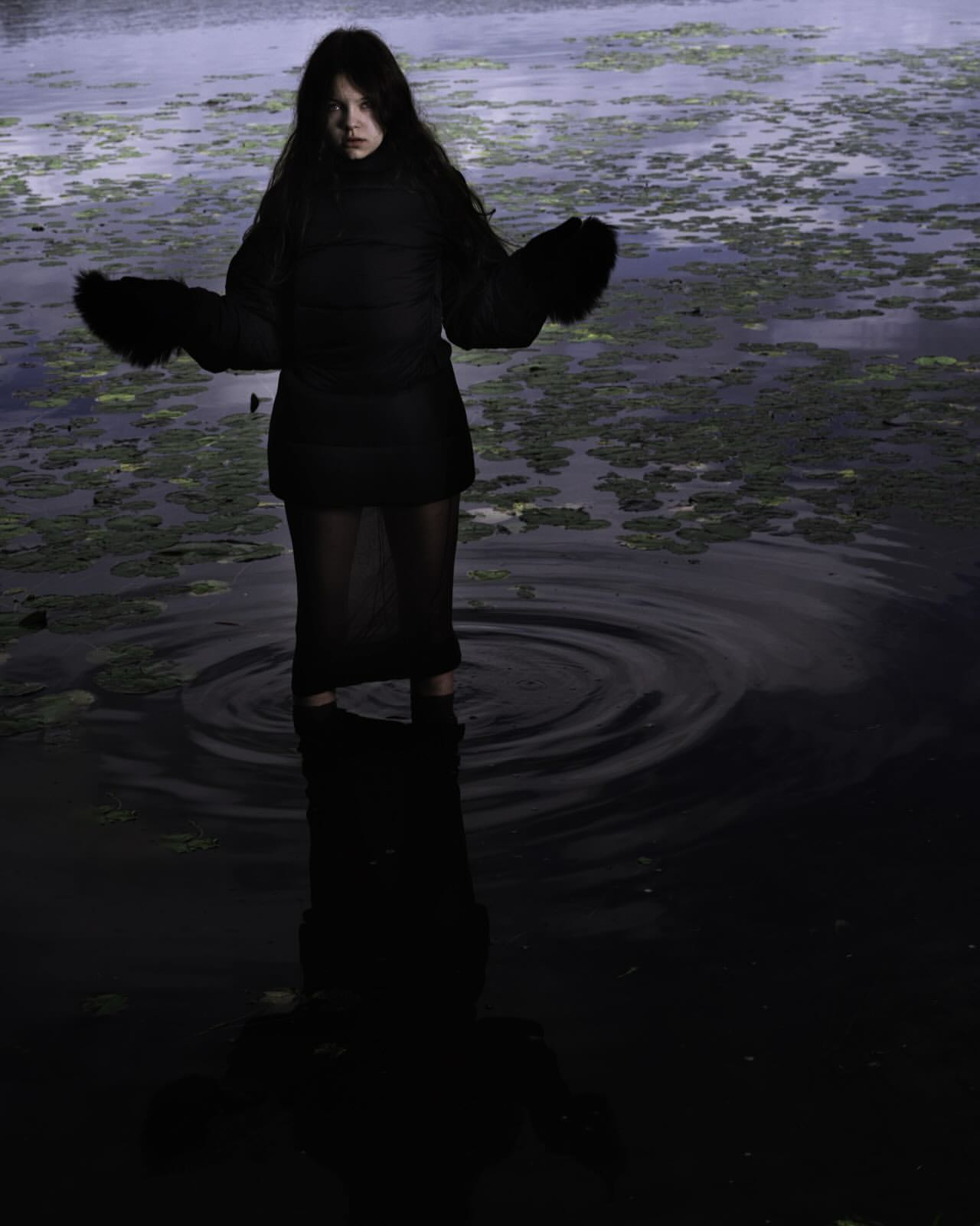 Woman in a black jacket and skirt standing in a swamp, surrounded by water and green plants. Dark and moody aesthetic.