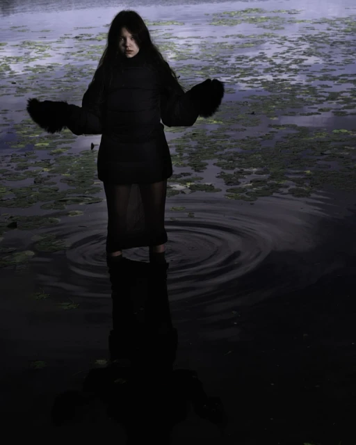 Woman in a black jacket and skirt standing in a swamp, surrounded by water and green plants. Dark and moody aesthetic.
