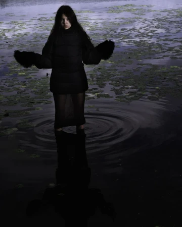 Woman in a black jacket and skirt standing in a swamp, surrounded by water and green plants. Dark and moody aesthetic.
