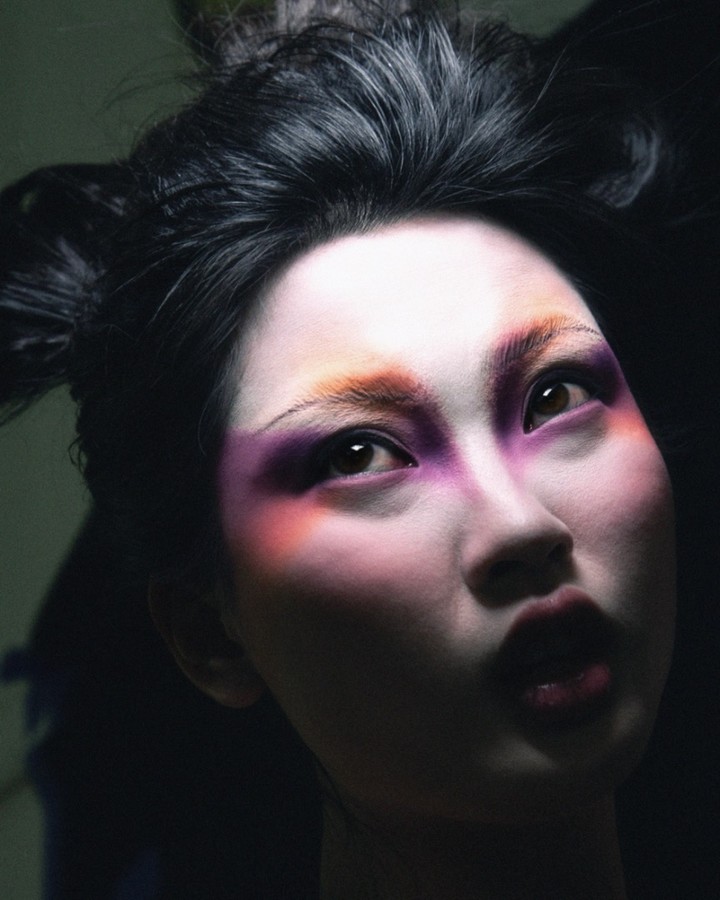 Dramatic close-up of an Asian woman with artistic makeup featuring vibrant purple and orange hues, set against a dark background.