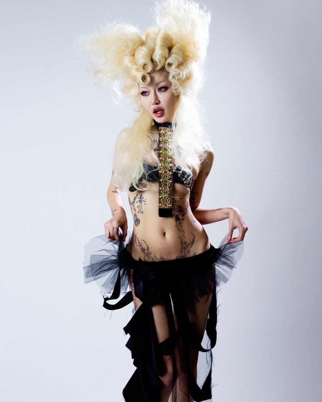 Striking full shot of a slender adult woman with elaborate blonde hair and black lingerie, posing in a photo studio against a white backdrop.