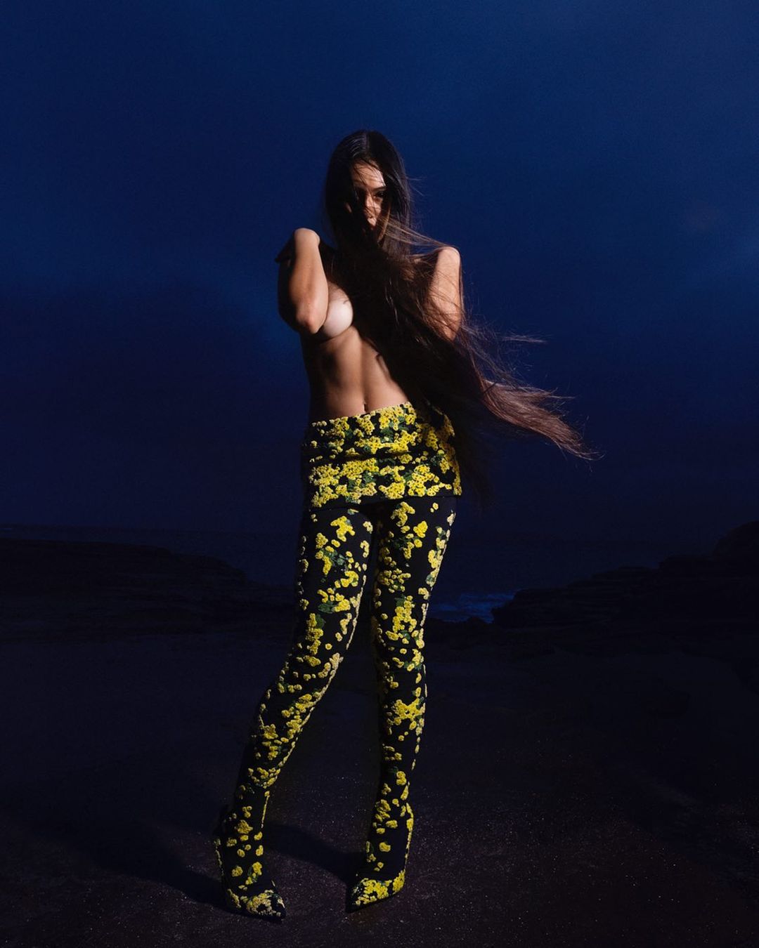 Woman with long hair poses in floral patterned pants against a dark blue background, exhibiting fashion and beauty elements in a striking portrait.