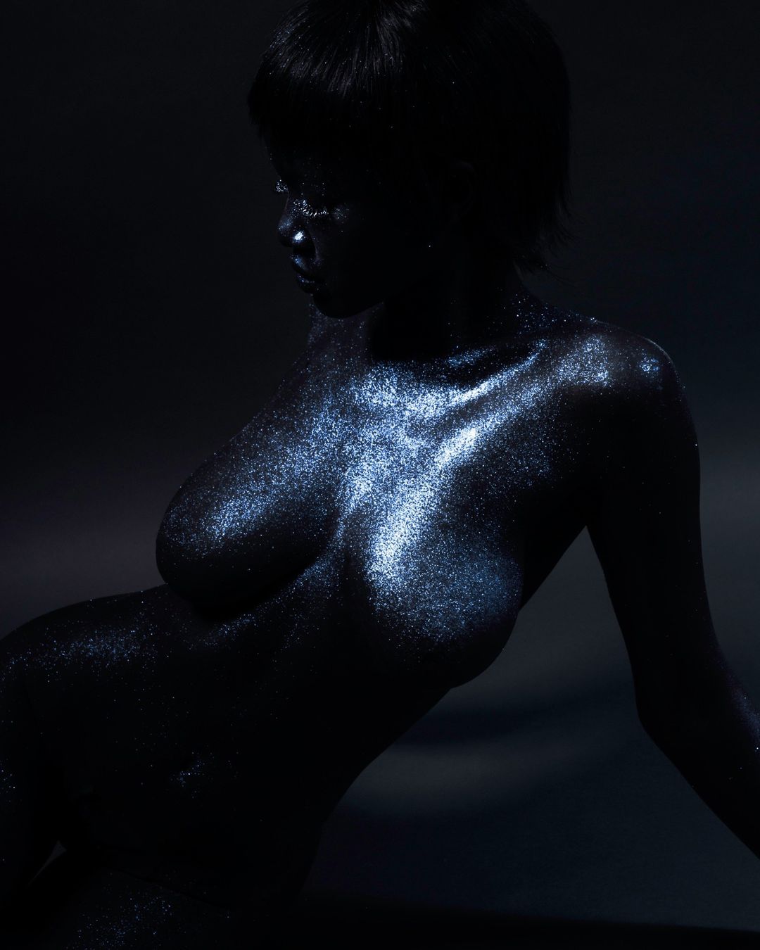 Shimmering nude adult female body covered in glitter, reclining pose, low key lighting, dark background, beauty and fashion editorial.