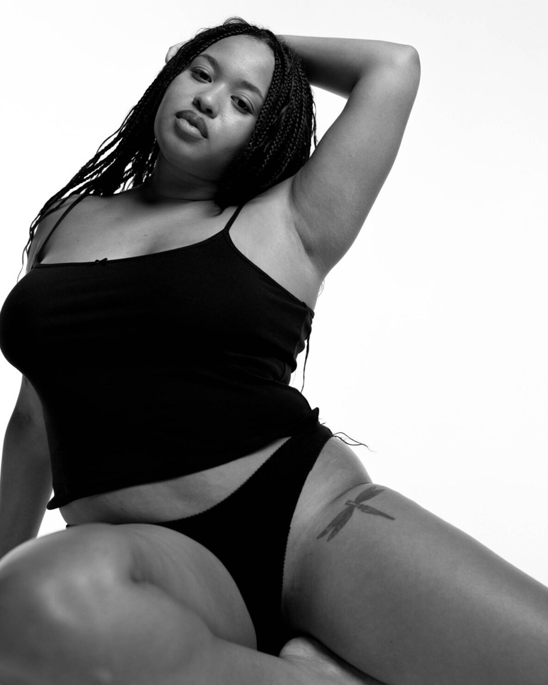 Monochrome portrait of a Black woman with braids, wearing a black top and underwear, posing in a photo studio against a white background.