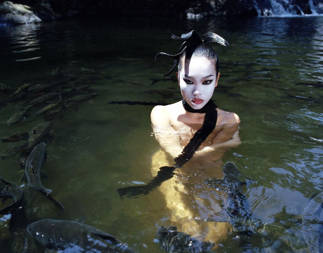 Artistic shot of a woman standing in a river surrounded by fish, wearing pale makeup and black fabric on her head and neck.