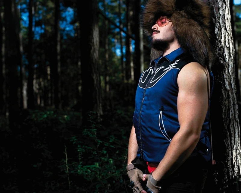 A man in a fur hat and vest stands near a tree in a forest setting. He wears sunglasses and gloves with a beard and mustache.