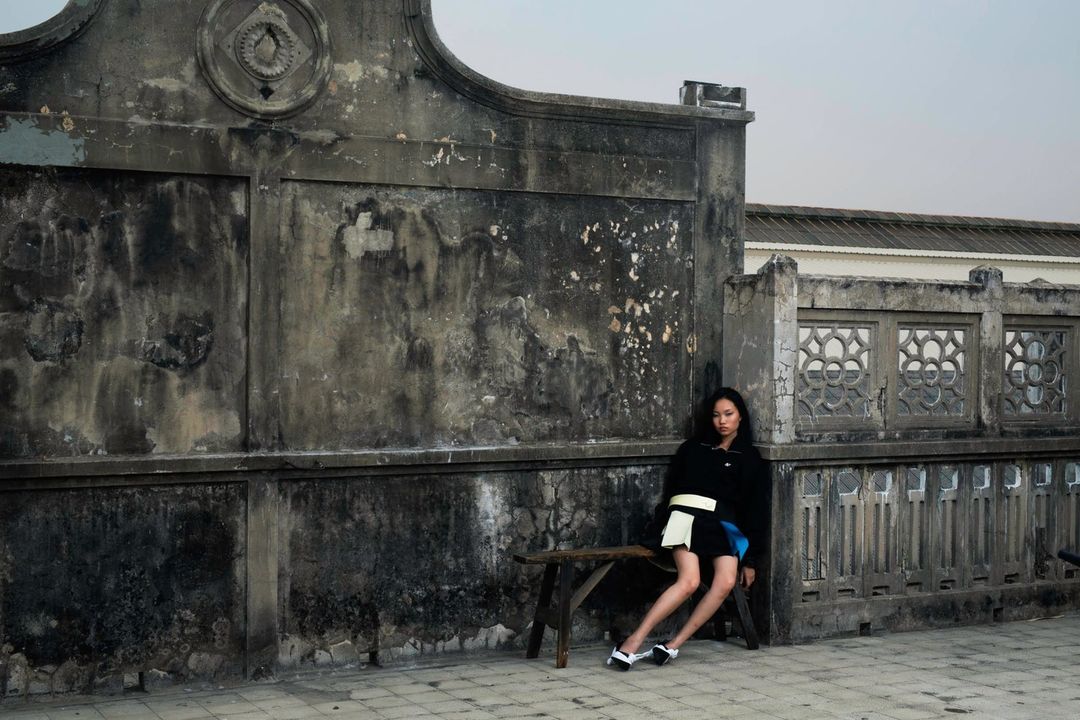 Fashion portrait of an Asian woman in a black jacket and blue skirt sitting on a bench in front of an old concrete building outdoors.