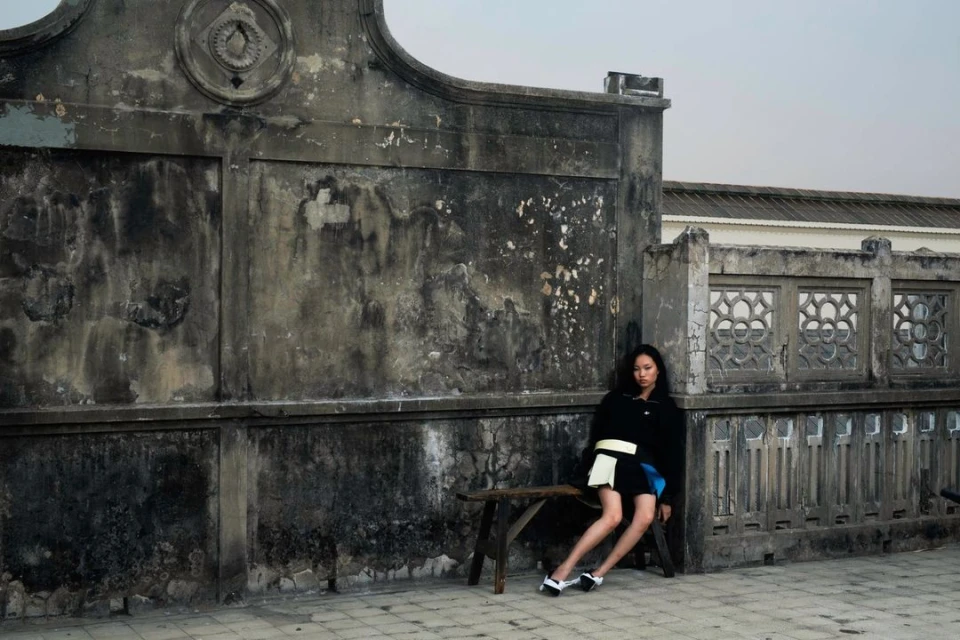 Fashion portrait of an Asian woman in a black jacket and blue skirt sitting on a bench in front of an old concrete building outdoors.