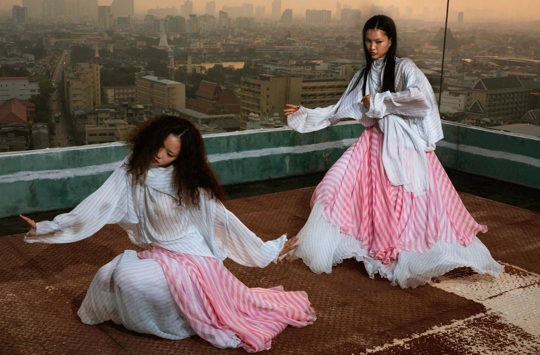 Two women in flowing striped dresses pose on a rooftop overlooking a cityscape at sunset, creating a fashion editorial-style image.