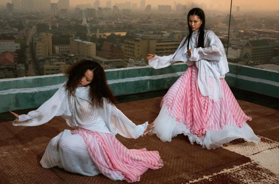 Two women in flowing striped dresses pose on a rooftop overlooking a cityscape at sunset, creating a fashion editorial-style image.